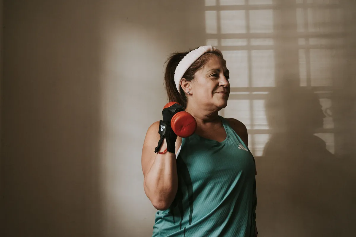 A woman in her 50s holding a dumbbell at her shoulder while training at home by a sunny window