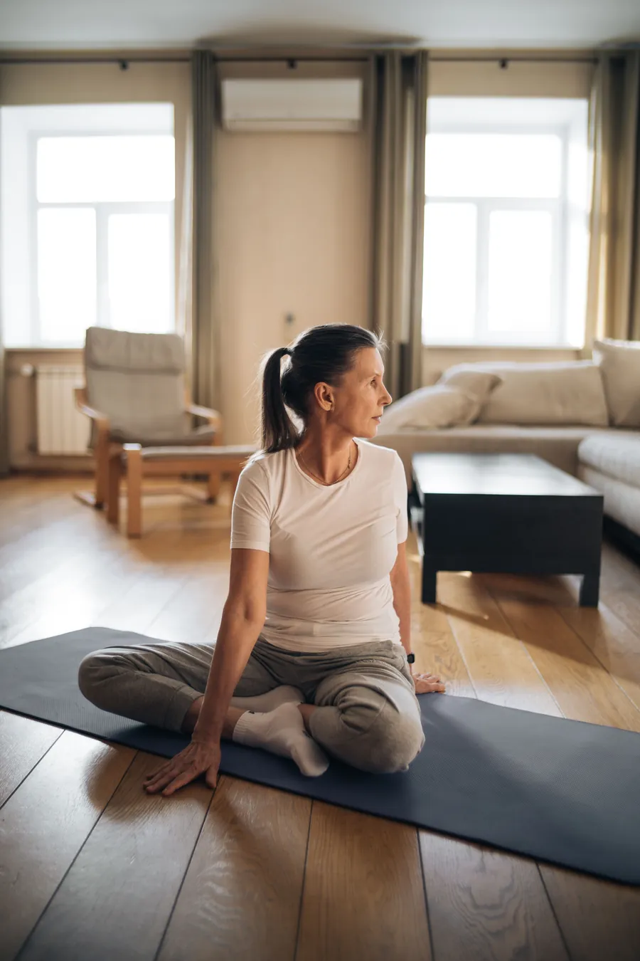 A woman in her 60s sitting on an exercise mat in her living room, stretching