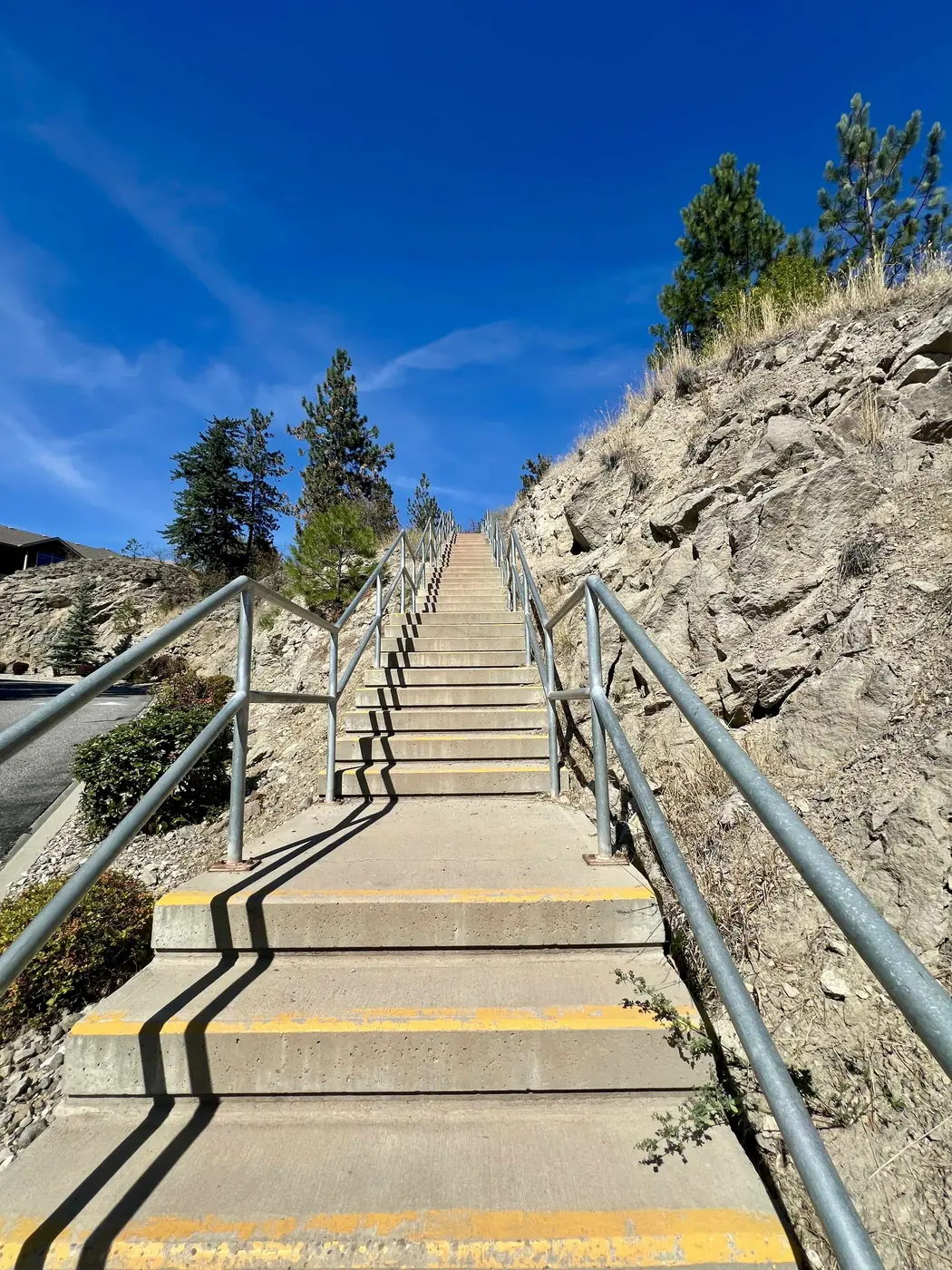 A long outdoor concrete staircase with metal handrails climbing up a hillside under a bright blue sky
