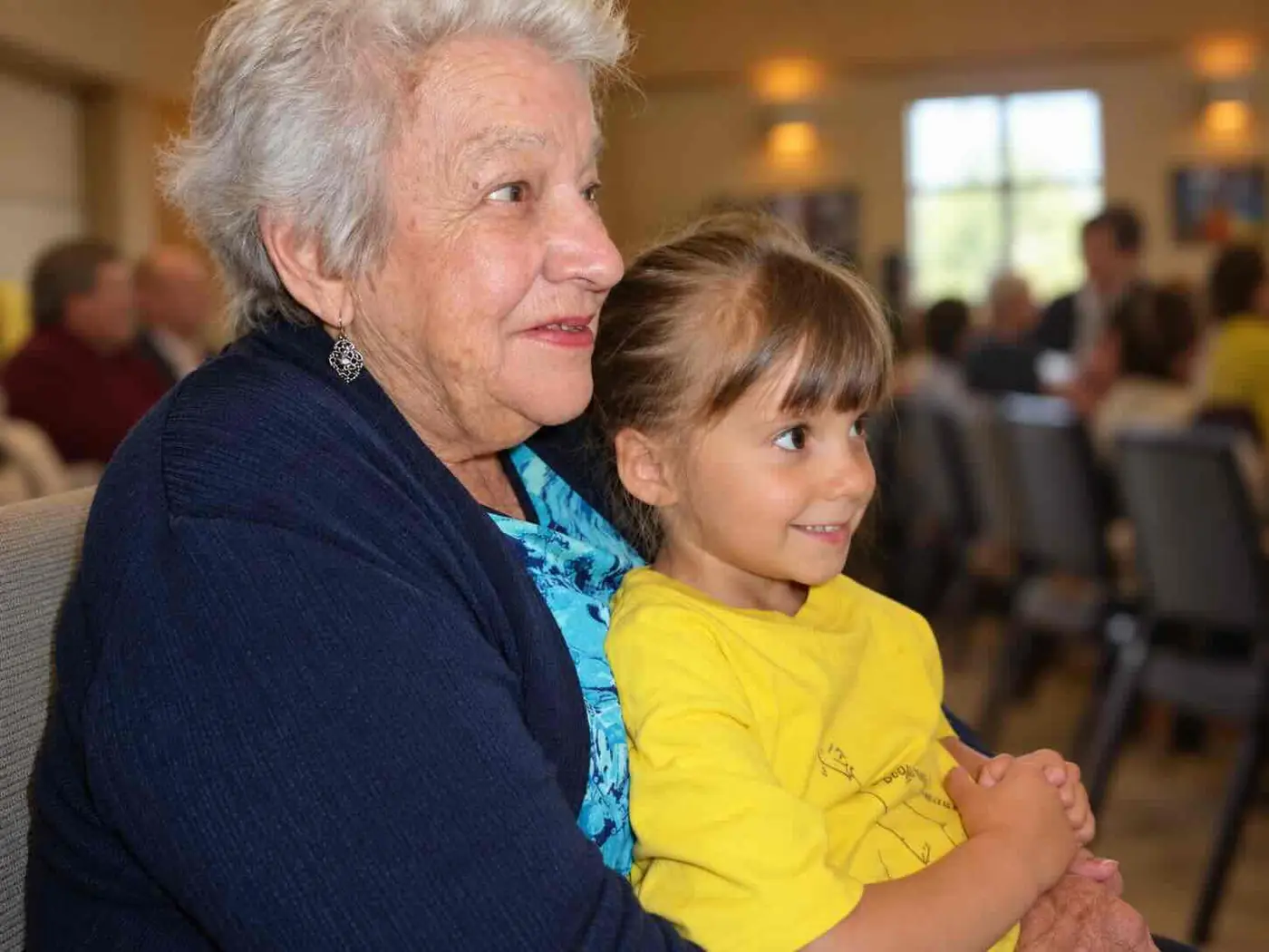 An older grandmother smiling and holding her young granddaughter seated together on a chair
