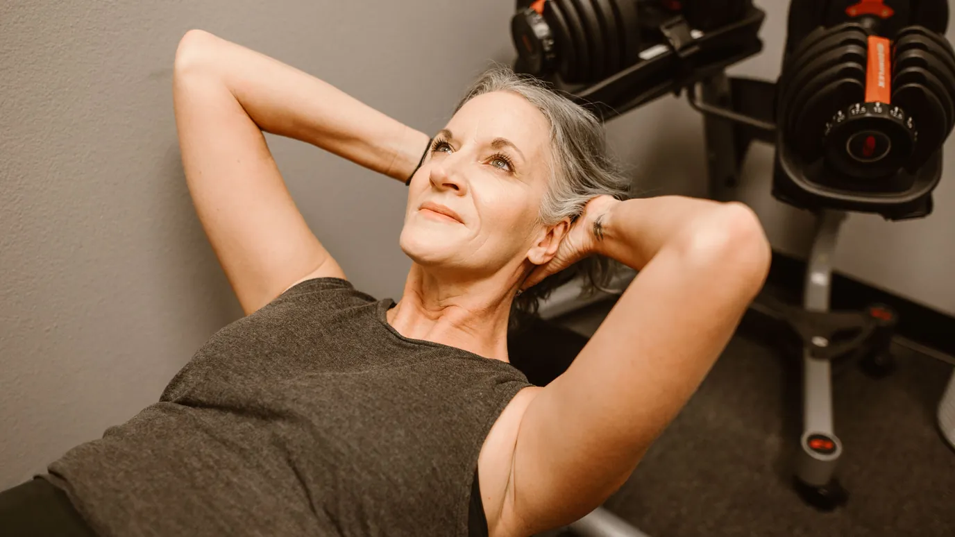 A woman with grey hair doing a crunch on a weight bench at home, with adjustable dumbbells on a rack beside her