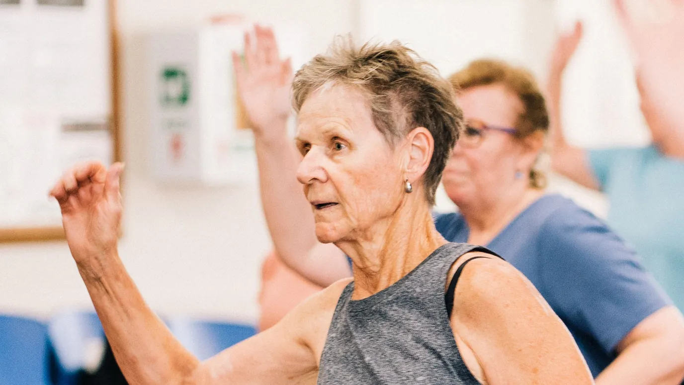 A woman with short grey hair dancing with her arms raised in a group exercise class