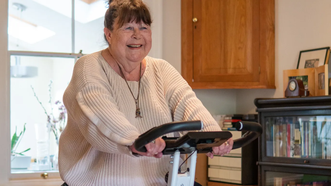 A smiling older woman in a knit sweater riding a folding exercise bike in her living room